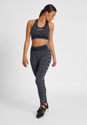 Kvinde i sort og grå mønstret sports-bh og leggings, der kigger mod højre, stående på ét ben i sorte sneakers mod en ensfarvet hvid baggrund.