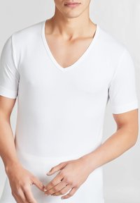 Maglietta bianca a maniche corte con scollatura a V realizzata in tessuto liscio. Presenta un design aderente e cuciture sulle spalle discrete. Mani posizionate sui fianchi.