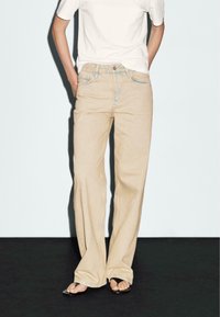 Jeans beige a gamba larga con vita alta, caratterizzati da cuciture chiare e chiusura a bottoni. Abbinati a una t-shirt bianca semplice e sandali.