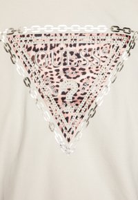 Logo en forme de triangle avec un fond en imprimé léopard, texte "GUESS USA", clous en argent et bordure en chaîne argentée sur un tissu beige.