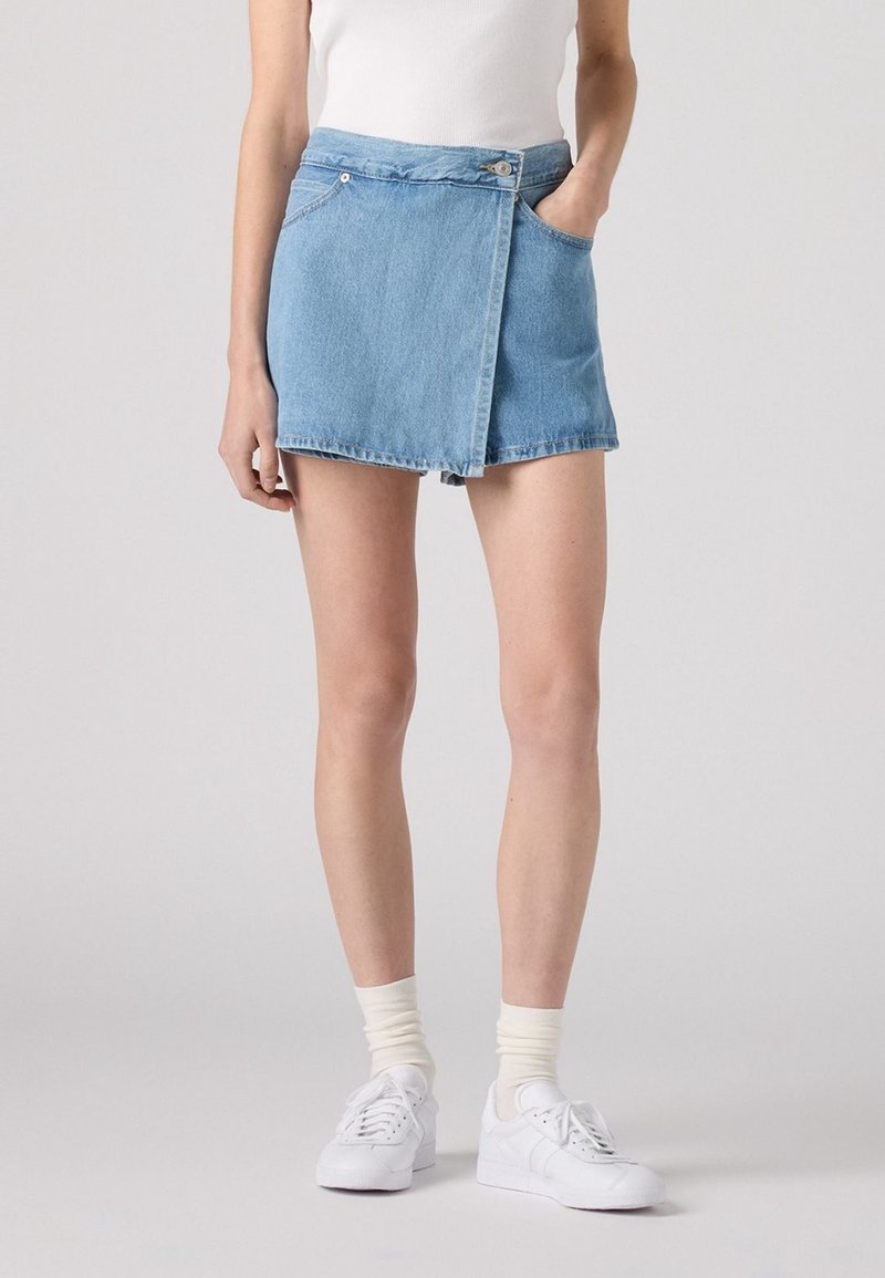 Jupe en denim mini de couleur bleu clair, avec un design enroulé, poche avant et fermeture à bouton. Associée à des baskets blanches et des chaussettes à côtes.