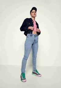 Jovem a usar um gorro preto, uma jaqueta de ganga escura, um top curto rosa, uns jeans claros e sapatilhas cor-de-rosa, verdes e pretas, a mostrar uma postura confiante.