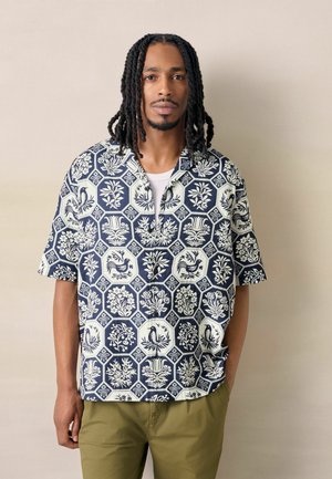 Mann mit schulterlangen Dreadlocks trägt ein marineblau-weiß gemustertes kurzärmeliges Hemd und olivgrüne Hose und steht vor einer schlichten Wand.