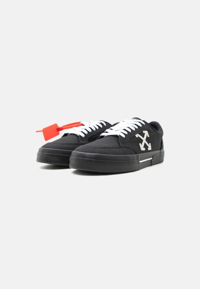 Baskets basses noires avec des lacets blancs, logo flèches blanches brodées sur les côtés et une étiquette rouge attachée à la chaussure gauche.