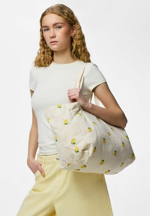 Noor naine lokkis blondide juustega kannab valget särki ja kollaseid pükse, kandes valget kandekotti kollase sidruni mustriga.