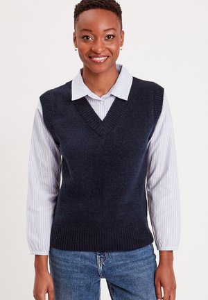 Gilet senza maniche di colore blu navy in morbido tessuto a maglia con scollatura a V e bordi a coste, indossato sopra una camicia a righe blu chiaro.