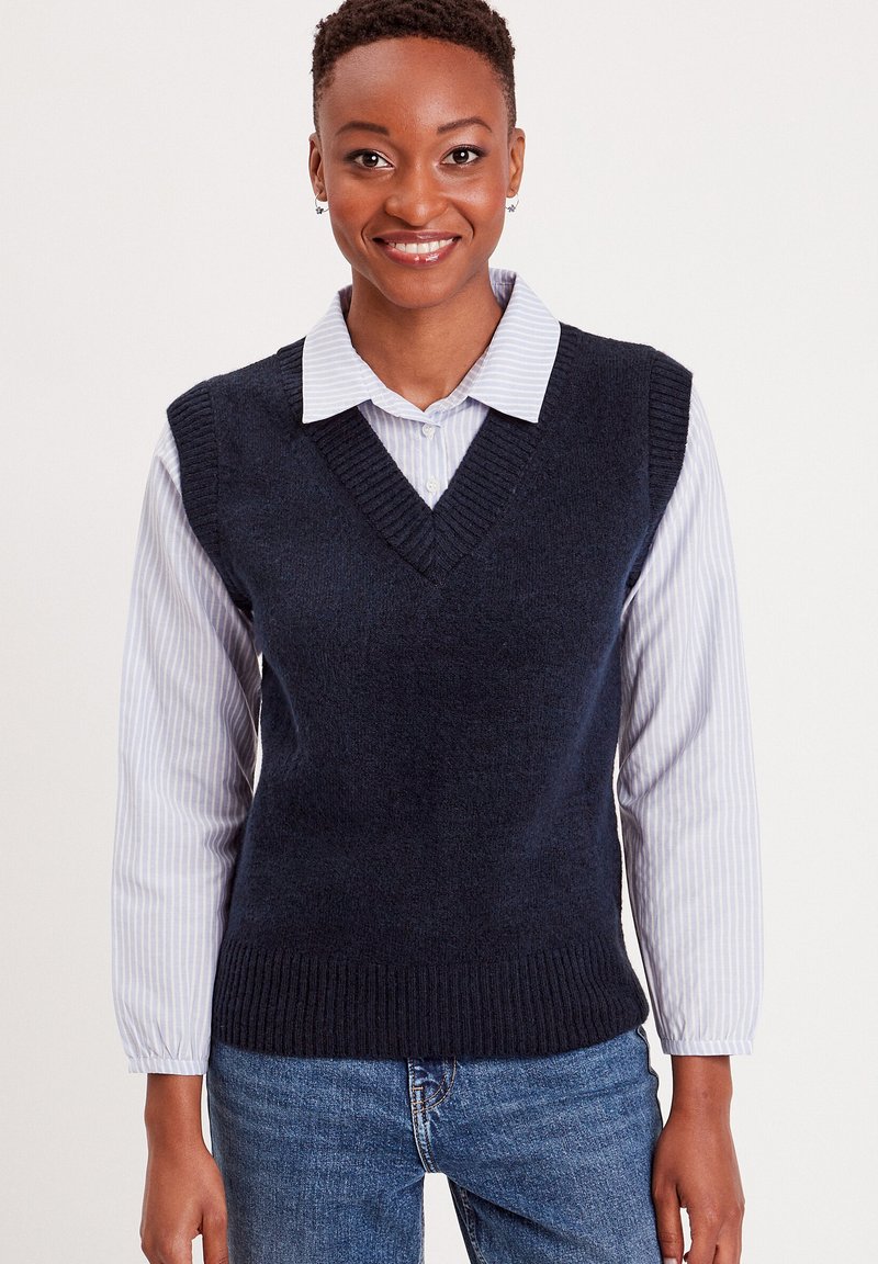 Marineblå ærmeløs sweater vest i blødt strikstof med V-hals og ribkanter, båret over en lyseblå stribet skjorte.