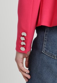 Manche de blouse rose vif avec quatre boutons ronds en argent. La manche a une texture lisse, associée à un jean en denim bleu foncé.