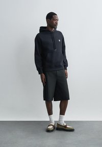 Haut à capuche en coton noir avec poche avant et cordons de serrage, associé à un short foncé. Des chaussettes blanches et des chaussures bicolores complètent la tenue.