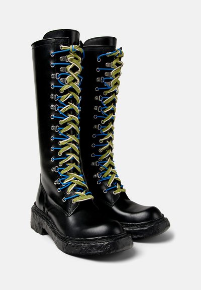 Botas de cuero negras hasta la rodilla con una suela gruesa y texturizada. Presentan cordones multicolores en azul y verde y ojales plateados en la parte delantera.