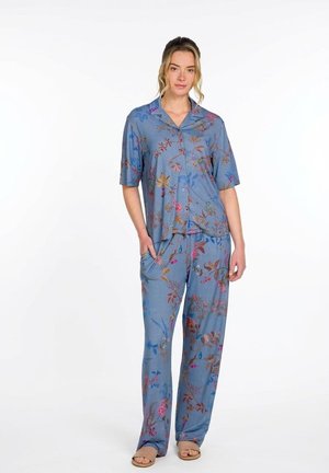 Frau trägt ein blaues, geblümtes, kurzärmeliges Hemd mit Knöpfen und passende, locker sitzende Hosen mit Taschen, steht mit einer Hand in der Tasche und trägt Sandalen.