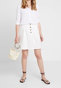 Valkoinen nappipaita, jonka hihat on kääritty, yhdistetty valkoiseen hameeseen, jossa on kultaiset napit. Läpinäkyvä käsilaukku ja mustat remmisandaalit täydentävät asua.