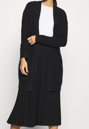 Personne portant un cardigan noir duveteux ouvert sur le devant, par-dessus une chemise blanche et une jupe midi plissée noire.