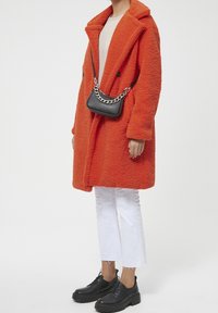 Personne portant un manteau texturé orange vif, un pantalon blanc court, des chaussures noires chunky et un sac bandoulière noir avec une chaîne en argent.