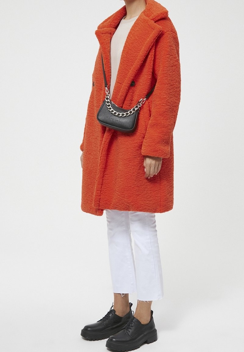 Personne portant un manteau texturé orange vif, un pantalon blanc court, des chaussures noires chunky et un sac bandoulière noir avec une chaîne en argent.