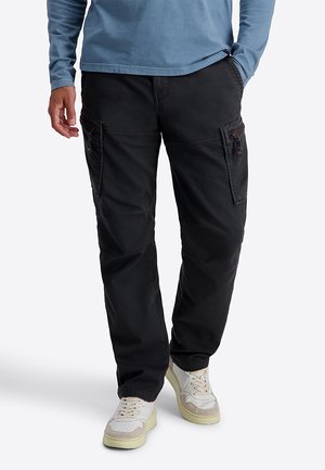 Mann trägt schwarze Cargohose und weiße Sneakers, eine Hand in der Tasche und ein teilweise sichtbares blaues Langarmhemd.