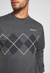 Grå sweatshirt med rund halsringning, prydd med ett geometriskt diamantmönster i marinblått och vitt, med Adidas-loggan på övre vänstra sidan.