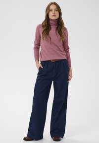 Roza turtleneck pulover v kombinaciji z modrimi širokimi hlačami. Pulover ima dolge rokave, medtem ko imajo hlače visok pas in nagubano sprednjo stran.