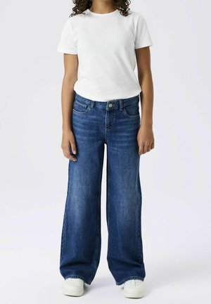 Osoba ubrana w prostą białą koszulkę z krótkim rękawem, szerokie niebieskie dżinsy i białe trampki, stojąca na tle jednolitego jasnego tła.