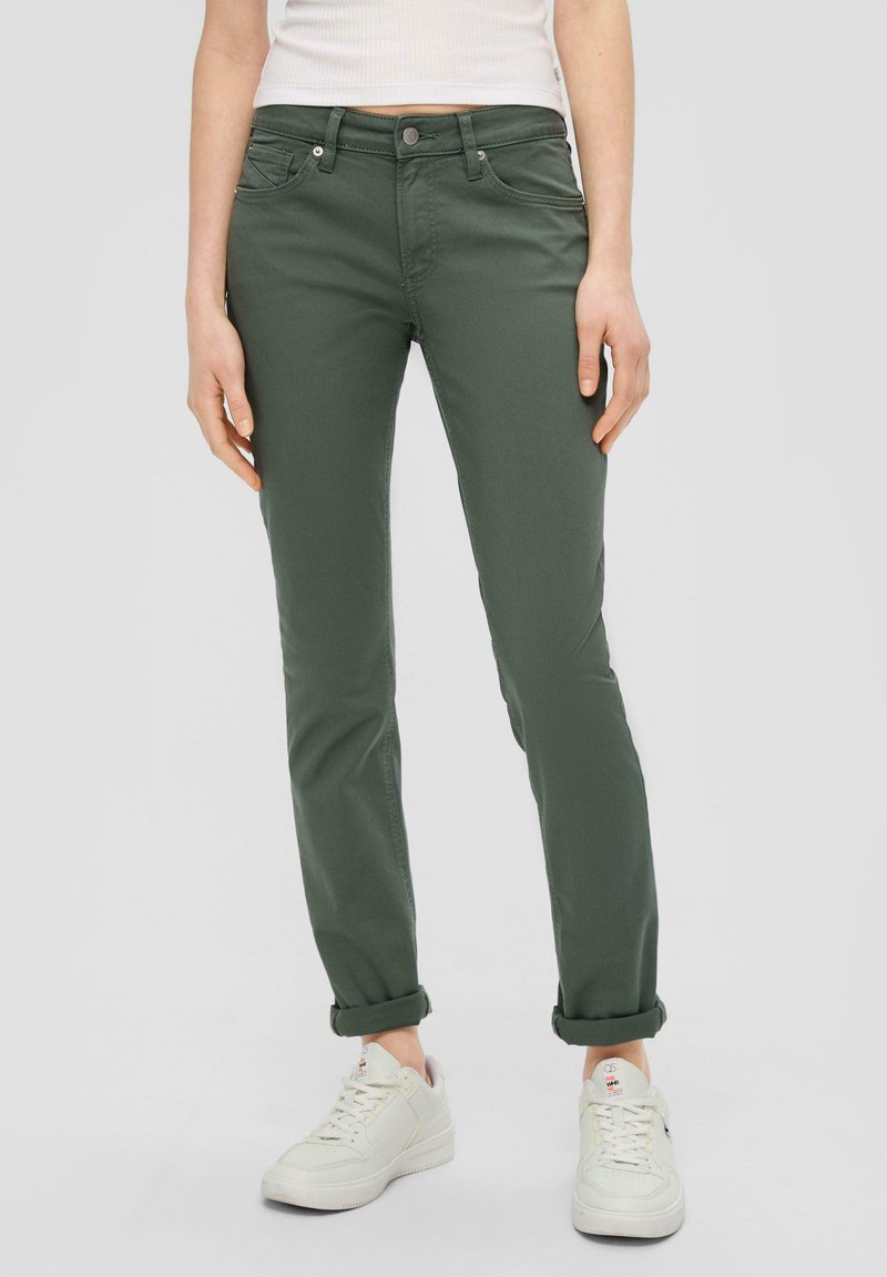 Femme portant un pantalon fitted vert olive roulé aux chevilles et des baskets blanches, debout devant un fond clair uni.