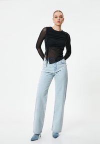 Top negro transparente de manga larga con detalles fruncidos en los laterales, combinado con jeans de pierna ancha de talle alto en azul claro y tacones azules de punta.