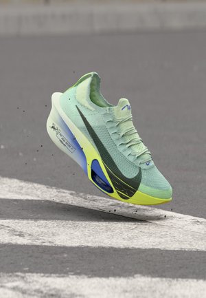Lichtgroene en gele Nike hardloopschoen met zwarte swoosh, zwevend boven asfalt met witte wegstrepen, waarbij de zool en veters zichtbaar zijn.