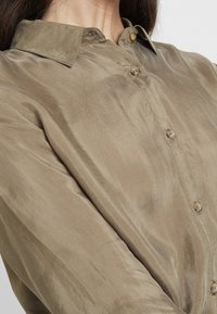 Olivově zelená, hedvábná košile na knoflíky s límečkem, s hladkým povrchem, pěti knoflíky a viditelným prošitím podél légne.