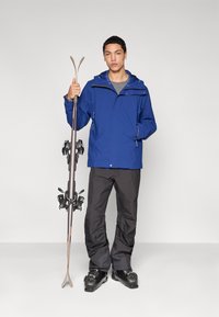 Blå vattentät jacka med huva, grå tröja under, svarta skidbyxor och skidbindningar. Modellen håller ett par skidor.