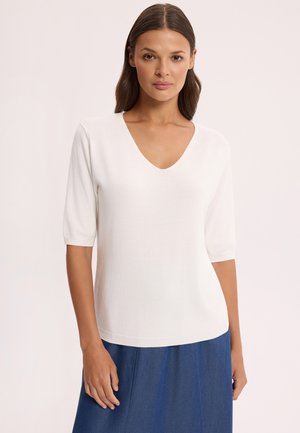 Kobieta o prostych brązowych włosach, ubrana w biały top z dekoltem w serek i rękawami do łokci oraz niebieską spódnicę, stojąca na jednolitym tle.