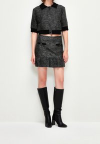 Veste en tweed noir et gris coupée avec un col, assortie d'une jupe coordonnée dotée de deux poches et d'un ourlet à volants. Bottes noires jusqu'aux genoux.