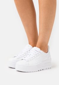 Bijele platform tenisice s glatkim materijalom sličnim koži, zaobljenim vrhom, debelim potplatom i dizajnom na vezanje s perforiranim detaljima.