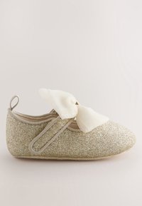 Une chaussure de bébé pailletée dorée avec une semelle souple et un nœud en tissu blanc sur la bride, présentée sur un fond clair.