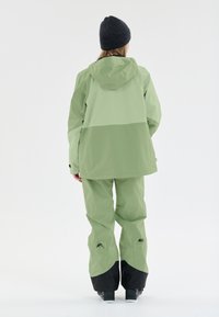 Vattentät, tvåtonad olivgrön jacka med huva och matchande byxor. Byxorna har svarta detaljer vid fållarna och blixtlåsen.