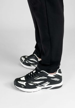 Persoon draagt zwarte en witte Dutch D sneakers en een zwarte broek, staande op een lichtgrijze vloer.