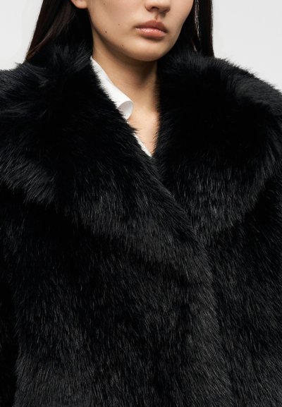 Μαύρο παλτό από ψεύτικη γούνα με φαρδύ, απαλό γιακά, που έχει απαλή, γυαλιστερή υφή και ελαφρώς κυματιστό σχέδιο, φορεμένο πάνω από ένα λευκό πουκάμισο.