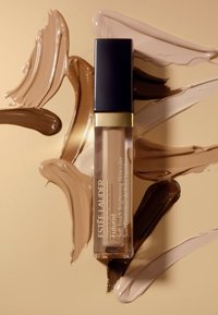 Estée Lauder Futurist concealer i en klar tube, med en sort kapsel og gyldne accenter, omgitt av forskjellige nyanser av krem- og brune produktprøver.