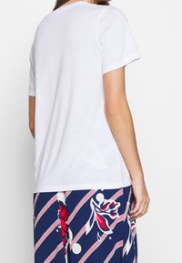 Camiseta blanca de corte relajado, con cuello redondo, mangas cortas y textura suave, vista desde la parte posterior junto a pantalones azules con estampado.