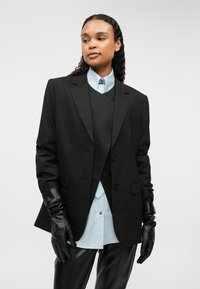 Čierny prispôsobený blejzer s dvoma gombíkmi, kombinovaný s svetlomodrou košeľou a čiernym vestom. Doplnené čiernymi koženými rukavicami a nohavicami.