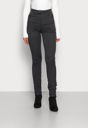 Femme portant un pantalon gris foncé ajusté, un haut blanc à manches longues côtelé, et des bottines noires à talons, debout devant un fond clair.