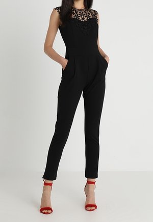 Zwarte jumpsuit met een sheer kanten lijfje met bloemenpatroon, een getailleerde pasvorm, zijzakken en smalle pijpen, gecombineerd met rode gespenhakken.