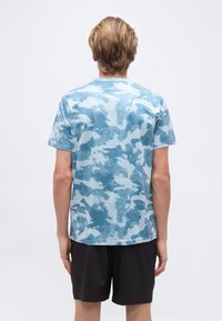 Άτομο με ξανθά μαλλιά που φορά μπλε και λευκό πουκάμισο κοντού μανικιού με σχέδια και μαύρο σορτσάκι, στέκεται γυρισμένο πλάτη σε απλό φόντο.