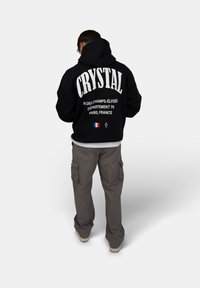 Felpa nera con la scritta "CRYSTAL" e dettagli di Parigi sul retro; indossata con pantaloni cargo grigi chiari e una maglietta bianca sotto.
