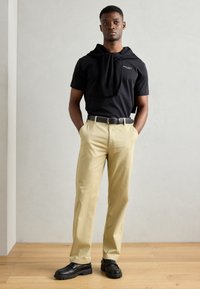 Zwarte korte mouwen shirt met een ronde hals en een trui om de schouders gedrapeerd, gecombineerd met lichtbeige broeken en zwarte instapschoenen.