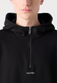 Melna aizdare ar rāvējslēdzēju, izgatavota no mīksta auduma. Satur priekšējo rāvējslēdzēju un kapuci. Iekļauj nelielu balto "Calvin Klein" logotipu uz krūtīm.
