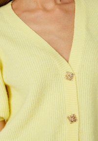 Kollane kootud kardigan V-kaelusega, kolme dekoratiivse kullatud ruudukujulise nupuga, ribakujulise tekstuuriga ja lühikeste varrukatega.