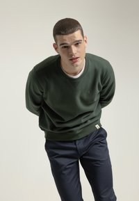 Sudadera de algodón verde con cuello redondo y puños de canalé, combinada con pantalones azul marino. Presenta un pequeño detalle de logo en el pecho.