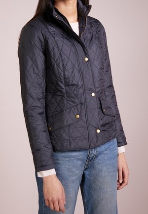 Femme portant une veste matelassée bleu marine avec des boutons dorés, une fermeture éclair, des poches à rabat et un jean bleu clair sur un fond uni.