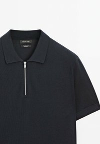 Černé polo tričko ze 100% merino vlny, s zipovým výstřihem, krátkými rukávy a hladkou texturou. Viditelné etikety uvnitř.