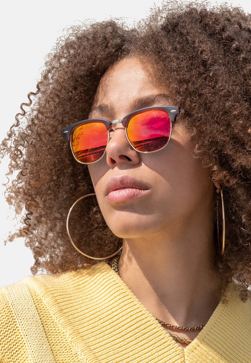 Reflective orange sunglasses with a black metal frame, featuring round lenses and a silver accent bridge. Worn with a yellow knit top.