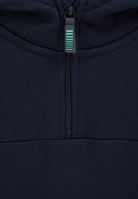 Tumši zils džemperis ar rāvējslēdzēju un vienmērīgu tekstūru. Rāvējslēdzēja vilcējā ir ekrāns kontrastējošā zaļā vertikālā svītrā. Minimālistisks dizains un šuves.